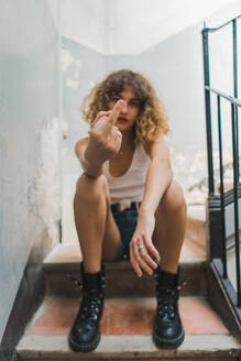 Junge lockige Frau in brutalen Stiefeln und Shorts sitzt auf einer schäbigen Treppe und zeigt Mittelfinger - ADSF00613