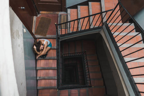 Blick von oben auf eine lockige Frau in kurzen Hosen, die auf einer schäbigen Treppe im Flur sitzt und nach oben schaut - ADSF00614