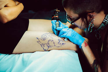 Side view of trendy female in latex gloves using professional tool to make tattoo on leg of anonymous client - ADSF00619