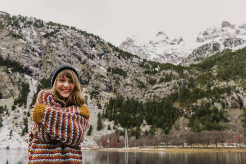 Attraktive junge Frau in Pullover und Hut mit gekreuzten Händen in der Nähe von Hügeln auf unscharfem Hintergrund in den Pyrenäen, lizenzfreies Stockfoto