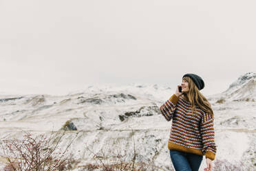 Lächelnde junge Frau in Pullover und Hut, die mit dem Handy telefoniert und in der Nähe von verschneiten Hügeln in den Pyrenäen wegschaut - ADSF00663