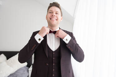 Young handsome smiling man in black costume standing in bedroom and putting on bow tie - ADSF00669