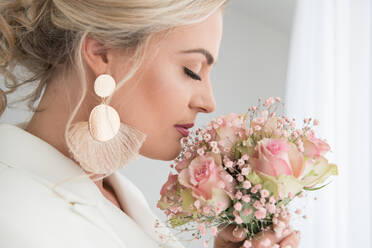 Schöne erwachsene blonde Frau trägt eleganten Brautanzug und Ohrringe riechen zarten Blumenstrauß im Tageslicht - ADSF00682