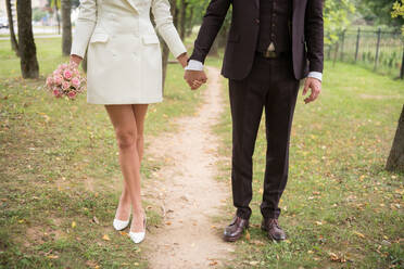 Gesichtslose Aufnahme eines eleganten, trendigen Paares, das sich an den Händen hält und auf einem Weg in einem grünen Park steht - ADSF00683