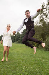 Trendy erwachsenen Bräutigam und Braut in eleganten Anzügen halten die Hände und springen mit Aufregung auf der grünen Wiese - ADSF00686
