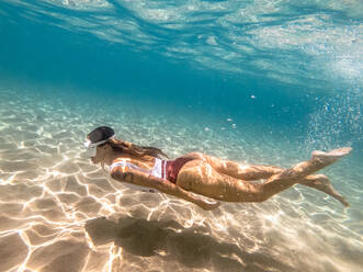 Seitenansicht von schönen fit weiblich mit Tätowierung auf Arm in Badeanzug und Tauchmaske Schwimmen unter Wasser auf sandigen Meeresboden - ADSF00780