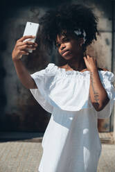 Stylish young African-American woman in white dress using phone and taking selfie on street in sunlight - ADSF00797