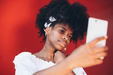 Stilvolle schwarze Frau mit Afro-Frisur und mit Piercing unter Selfie mit Smartphone auf helle rote Wand - ADSF00806