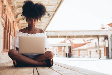 Hübsche moderne afroamerikanische Frau in weißem Kleid sitzt mit gekreuzten Beinen auf dem Bürgersteig und benutzt einen Laptop im Sonnenlicht - ADSF00815