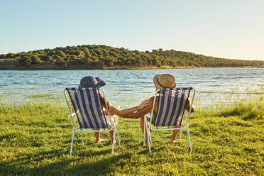 Back view young couple in hats holding hands and sitting on folding chairs on coast with green grass near water in summer - ADSF00827