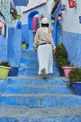 Rückenansicht einer Dame in Rock und Hut, die eine blaue Treppe in der Nähe alter Häuser in Marrakesch, Marokko, hinaufgeht - ADSF00862