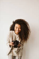 Junge Afroamerikanerin in modischem Outfit, die eine moderne Fotokamera hält und lacht, während sie an einer weißen Wand steht - ADSF00866