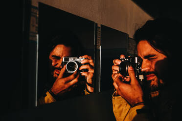 Erwachsener Mann mit Fotokamera, der durch einen Spiegel an der Wand in goldenem Licht auf die Kamera gerichtet ist - ADSF00875