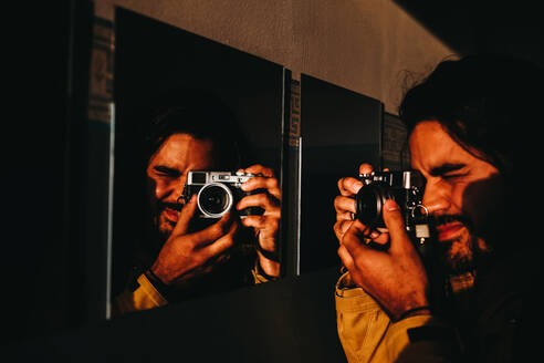 Adult man with photo camera focusing at camera through mirror on wall in golden light - ADSF00875