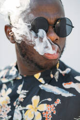 Black stylish man wearing sunglasses. He is vaping with an electronic cigarrete outdoor - ADSF00893