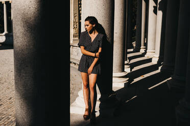 Wunderschöne junge Frau in stilvollem Kleid lehnt an einer Säule eines alten Gebäudes an einem sonnigen Tag in Madrid, Spanien - ADSF00903