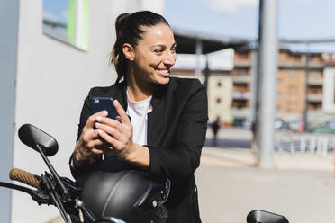 Lächelnde Frau sitzt auf einem Motorrad und benutzt ein Smartphone - FMOF01018