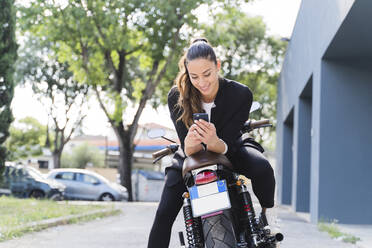 Lächelnde Frau sitzt auf einem Motorrad und benutzt ein Smartphone - FMOF01022