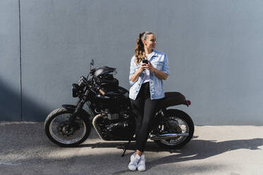 Smiling woman sitting on motorbike and using smartphone - FMOF01024