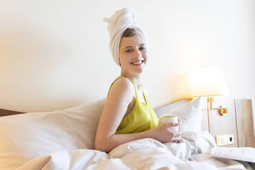 Lächelnde schöne Frau, die ein Handtuch trägt, während sie einen Kaffee auf dem Bett zu Hause genießt - TAMF02478