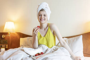Lächelnde Frau mit Handtuch, die zu Hause auf dem Bett frische Erdbeeren genießt - TAMF02479