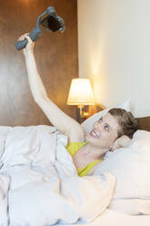 Glückliche schöne Frau, die ein Selfie macht, während sie auf dem Bett zu Hause liegt - TAMF02486