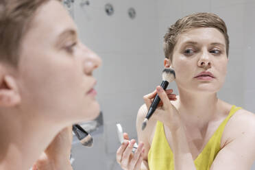 Kurzhaarige Frau trägt Make-up mit einem Pinsel auf, während sie ihr Spiegelbild im Badezimmer betrachtet - TAMF02488