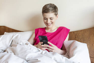 Lächelnde schöne Frau mit Smartphone beim Entspannen auf dem Bett zu Hause - TAMF02489