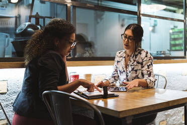 Young businesswoman pointing at diary while planning with female entrepreneur in coffee shop - MTBF00524