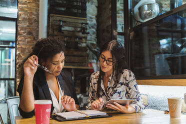 Multi-ethnic female colleagues discussing over digital tablet and diary while sitting at coffee shop during meeting - MTBF00526