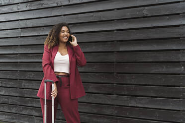 Lächelnde, schöne Unternehmerin, die mit einem Handy spricht, während sie mit Gepäck vor einer schwarzen Holzwand steht - MTBF00529