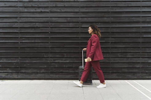 Young female professional walking with luggage on footpath by black wooden wall in city - MTBF00530