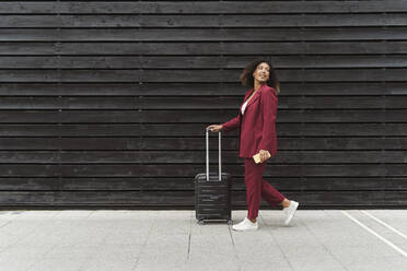Junge Geschäftsfrau blickt zurück, während sie mit Gepäck auf dem Fußweg an einer schwarzen Holzwand in der Stadt spazieren geht - MTBF00531