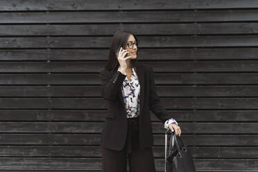 Beautiful female entrepreneur looking away while talking on mobile phone against black wooden wall - MTBF00533