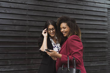 Happy female entrepreneur sharing smart phone while standing against black wall - MTBF00536