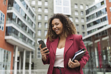 Smiling young businesswoman using smart phone while standing against building in city - MTBF00542