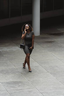 Fröhliche junge Geschäftsfrau, die über ihr Smartphone spricht, während sie auf einem Fußweg in der Stadt spazieren geht - MTBF00546