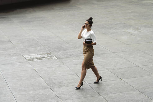 Junge Geschäftsfrau, die beim Gehen auf einem Fußweg in der Stadt mit ihrem Mobiltelefon spricht - MTBF00547