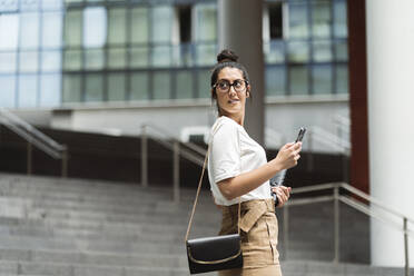 Schöne junge Geschäftsfrau schaut über die Schulter und hält ein Smartphone in der Stadt - MTBF00550