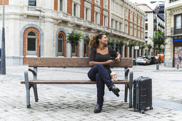 Lächelnde junge Geschäftsfrau in eleganter Freizeitkleidung sitzt auf einer Bank neben einem Gepäckstück - MTBF00551