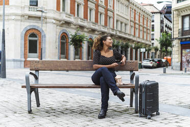 Smiling young businesswoman in smart casual sitting on bench by luggage - MTBF00551