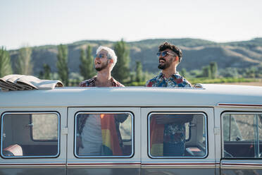 Zwei junge Männer umarmen sich und halten eine wehende LGBT-Flagge über den Köpfen, während sie in einem Retro-Van mit geöffnetem Dach in der Natur stehen - ADSF01011