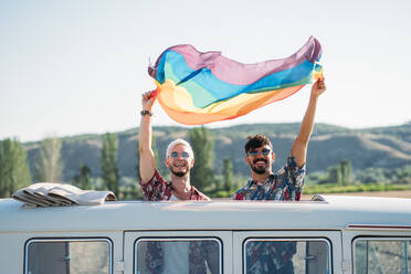 Zwei junge Männer umarmen sich und halten eine wehende LGBT-Flagge über den Köpfen, während sie in einem Retro-Van mit geöffnetem Dach in der Natur stehen - ADSF01012