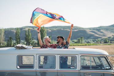 Zwei junge Männer umarmen sich und halten eine wehende LGBT-Flagge über den Köpfen, während sie in einem Retro-Van mit geöffnetem Dach in der Natur stehen - ADSF01013