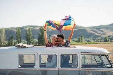 Zwei gutaussehende Männer küssen sich und halten eine helle LGBT-Fahne, während sie in einem Van mit geöffnetem Dach auf dem Lande an einem sonnigen Tag stehen - ADSF01014