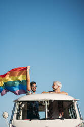 Zwei junge Männer umarmen sich und halten eine wehende LGBT-Flagge über den Köpfen, während sie in einem Retro-Van mit geöffnetem Dach in der Natur stehen - ADSF01019