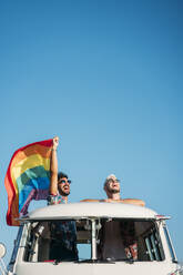 Zwei junge Männer umarmen sich und halten eine wehende LGBT-Flagge über den Köpfen, während sie in einem Retro-Van mit geöffnetem Dach in der Natur stehen - ADSF01020