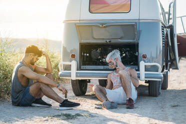 Two young men with repair tools sitting on ground near broken retro van and relaxing while travelling in countryside together - ADSF01024