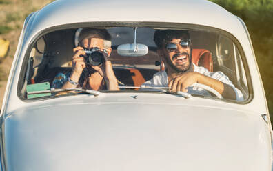 Two cheerful young guys with camera driving retro car and making pictures while travelling in countryside together - ADSF01026