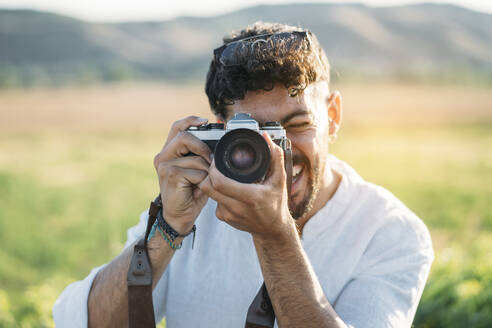 Gut aussehender junger Mann mit Sonnenbrille, der fröhlich lächelt und eine Retro-Fotokamera in der Hand hält, während er vor einem unscharfen Hintergrund mit beeindruckender Landschaft steht - ADSF01028